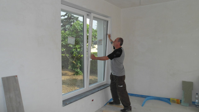 Vista interna di una stanza in ristrutturazione, con un operaio che collauda l’anta a ribalta di una finestra, attrezzi e materiali da lavoro sul pavimento